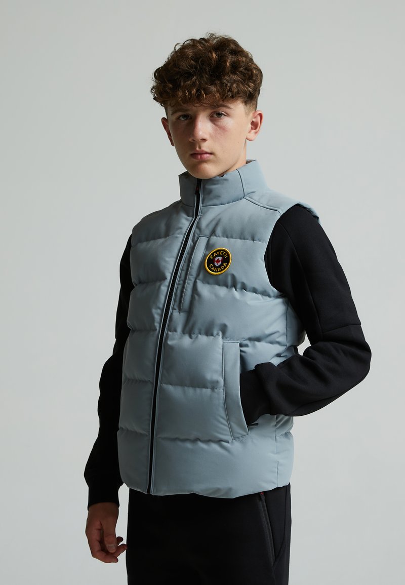 Gilet matelassé bleu clair avec col montant et fermeture éclair, arborant un patch logo rond, associé à un t-shirt à manches longues noir.