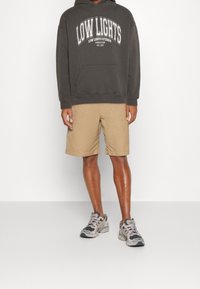 Una persona è in piedi rivolta in avanti, indossa un hoody scuro con la scritta "LOW LIGHTS" stampata su di esso, pantaloni corti khaki e scarpe da ginnastica. Lo sfondo è completamente bianco.