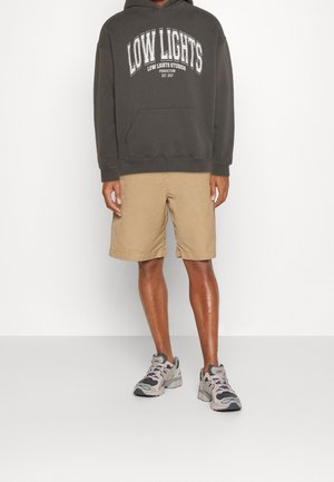En person står fremadskuende, iført en mørk hoodie med "LOW LIGHTS" trykt på, khaki kortbukser og sneakers. Baggrunden er ensfarvet hvid.