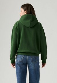 Sudadera verde con capucha de corte relajado, que presenta mangas largas y un bolsillo canguro, combinada con jeans de pierna ancha azules.