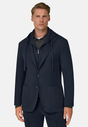 Homme portant un blazer bleu marine superposé à un sweat à capuche zippé et un polo gris foncé, debout devant un fond clair uni.