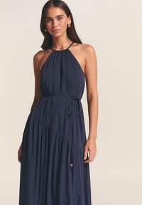 Marinblå halterneckklänning i lätt tyg, med rynkad halsringning, midjeband och vågigt, asymmetriskt fåll med ribbad struktur.