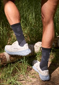 Weiße Trailrunning-Schuhe mit blauen Sohlen, strukturierter Oberseite mit Logo, kombiniert mit schwarzen knielangen Socken mit geometrischem Muster.