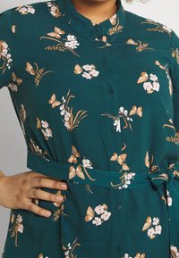 Teal jurk met knopen en een bloemen- en vlinderprint in beige en wit. Inclusief een bijpassende stoffen riem en korte mouwen.
