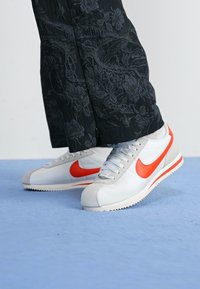 Baltos odinės sportinės bateliai su pilkais akcentais ir ryškiai oranžiniu "swoosh" logotipu. Turi tekstūruotą viršutinę dalį ir klasikę žemą formą.