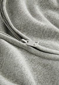 Grauer Stoff mit einer subtilen Textur, ausgestattet mit einem Metallreißverschluss und einem rechteckigen Ziehband, das entlang des Randes teilweise geöffnet ist.
