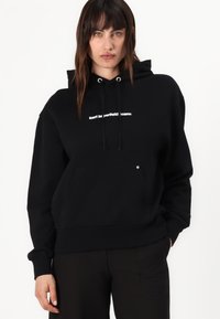 Felpa nera con cappuccio realizzata in tessuto morbido, dotata di tasca frontale a marsupio, cappuccio con cordino e logo bianco sul petto. Design semplice.