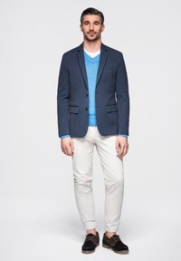 Homme debout portant un blazer bleu foncé, un pull bleu clair, un pantalon blanc et des chaussures noires sur un fond clair uni.