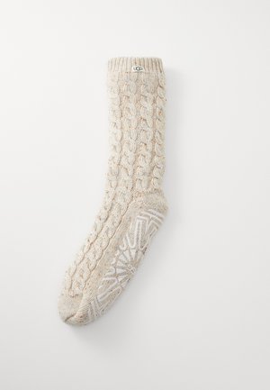 Cable-knit cream sock with a textured pattern, featuring a reinforced white rubber sole with geometric designs and a brand label at the top.