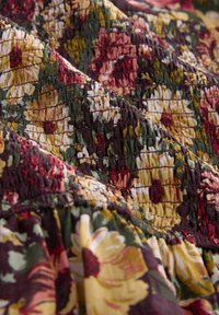 Mönstrad tyg med blommor i nyanser av grönt, bordeaux och gult. Har texturerad smock och gatherade kanter, vilket framhäver en livfull design.