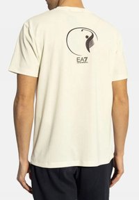 T-shirt color crema realizzato in tessuto morbido con scollatura rotonda. Presenta un logo grafico nero e un design sul retro. Maniche corte.