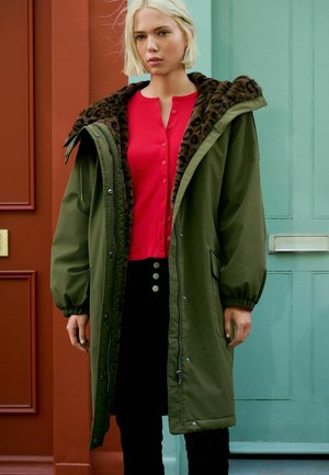 Blonde vrouw in lange groene parka met een voering van luipaardprint, rode getailleerde trui en zwarte broek, staand voor teal en rode deuren.