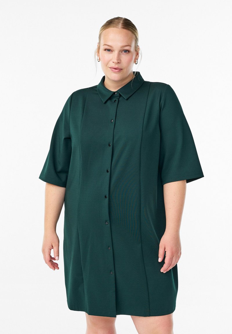 Eine Frau trägt ein dunkelgrünes, knöpfbares Hemdkleid mit Ärmeln bis zu den Ellenbogen und kleinen Creolen und steht vor einem weißen Hintergrund.