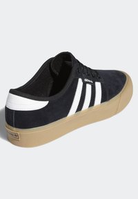 Svart mockasinsneaker med vita ränder, gummisula och snörningsdesign. Har en vadderad krage och adidas-logotyp på sidan och hälen.