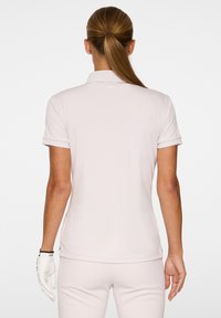 Jasnoróżowa koszulka polo z krótkim rękawem, elastyczny materiał, płaski kołnierzyk i subtelne logo z tyłu. Gładka tekstura i dopasowany krój.