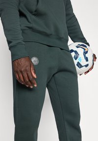 Mann i grønn treningsdrakt som holder en hvit og blå Nike fotball, Chelsea FC-logoen er synlig på buksene nær venstre lomme.
