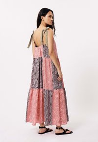 Robe maxi avec un patchwork de motifs floraux rose et noir, dos ouvert avec des bretelles à nouer, et une jupe à volants. Portée avec des sandales noires.