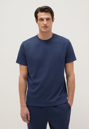 Uomo che indossa una t-shirt blu navy a manica corta con scollo rotondo e pantaloni blu navy abbinati, in piedi davanti a uno sfondo chiaro e uniforme.