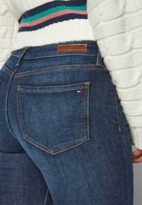 Detailní záběr na zadní kapsu modrých džín s malou vlajkou Tommy Hilfiger a hnědou koženou nášivkou značky, doplněný bílým pleteným svetr.