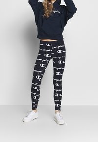 Un sweat à capuche bleu marine avec une poche avant, associé à des leggings ajustés ornés d'un texte "Champion" blanc répétitif. Des baskets blanches complètent la tenue.
