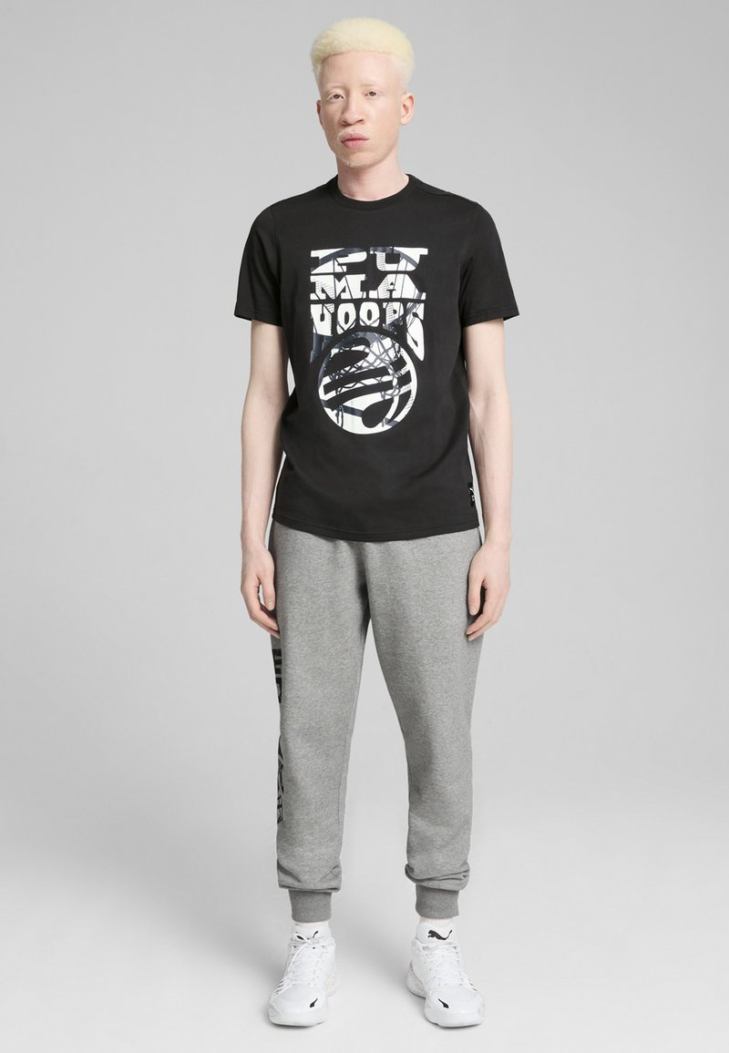Svart t-shirt med grafiskt tryck, grå sweatpants och vita sneakers. T-shirten har rund hals; sweatpants är elastiska vid muddarna.