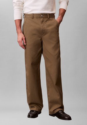 Mand iført brune bukser med lige ben, hvid langærmet skjorte og mørkebrune sko, stående med den ene hånd i lommen.