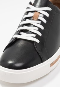 Baskets en cuir noir avec lacets blancs, semelle texturée et un accent beige sur le côté, présentant un bout arrondi et une finition lisse.