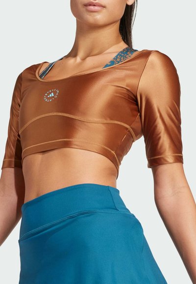 Hnědý crop top vyrobený z lesklé látky, s krátkými rukávy a modrými vzorovanými ramínky viditelnými pod ním. V kombinaci s jednobarevnou modrou sukní.