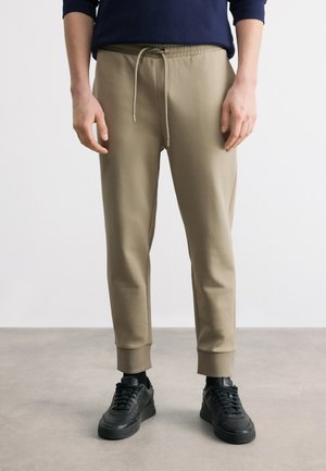 Uomo che indossa pantaloni da jogging beige con vita regolabile con coulisse, polsini a costine, calzini neri e sneakers nere su pavimento grigio con sfondo bianco.