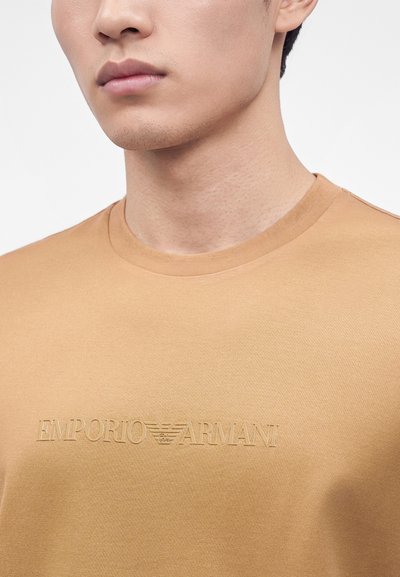 Νεαρός άνδρας φοράει μπεζ πουκάμισο Emporio Armani με ανάγλυφο λογότυπο στο στήθος, εμφανίζοντας το κάτω μέρος του προσώπου και τον λαιμό.
