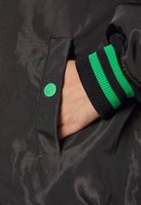 Svart jacktyg med en slät textur, med en grön tryckknapp och svart ribbad mudd med gröna ränder. Hand i fickan.