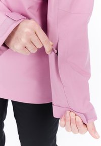 Person wearing a pink jacket adjusting a Velcro cuff and pulling a zipper on the sleeve, black pants visible below.