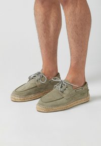 Espadrilles en toile vert olive avec une semelle en jute, bout rond et design à lacets. Présentent des coutures beige et un détail de marque sur le côté.