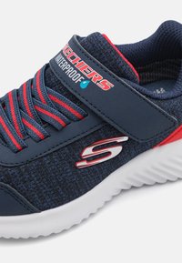 Sneaker imperméable bleu marine avec des accents rouges, tissu texturé, lacets élastiques et une sangle Velcro. Comprend une semelle en caoutchouc blanche et un détail de logo.