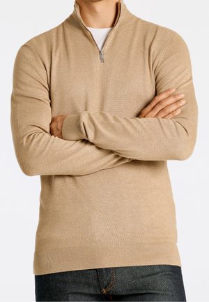 Homme portant un pull beige à fermeture éclair courte et un jean foncé, bras croisés, sur fond blanc uni, seul le torse et les bras sont visibles.
