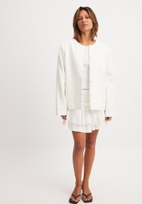 Witte katoenen blazer in een mix met een ronde hals, knopen aan de voorkant en lange mouwen, gecombineerd met een gelaagde witte minirok en zwarte sandalen.