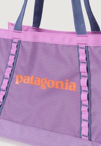 Tote taška Patagonia v purpurovej nylonovej farbe s ružovými akcentmi. Obsahuje modré rúčky, volánový detail a oranžové logo na prednej strane.