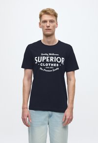 Navy blue short-sleeve t-shirt featuring white graphic print text. Simple round neck, soft fabric, and a relaxed fit. Paired with light blue jeans.