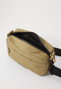 Borsa a tracolla in tessuto khaki con chiusura a zip, superficie testurizzata, tasca frontale, tracolla regolabile e etichetta con logo all'interno.