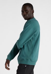 Sudadera azul verdosa con cuello redondo, mangas largas, puños y dobladillo acanalados. Presenta una textura suave y detalles en paneles laterales.