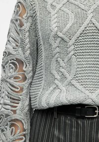 Pull en maille grise avec des motifs en relief, détail en dentelle transparente sur les manches et texture côtelée. Assorti à un pantalon rayé.