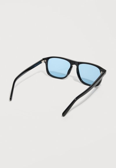 Μαύρα ορθογώνια γυαλιά ηλίου με φακούς μπλε απόχρωσης, ανοιχτά πάνω σε λευκή επιφάνεια.