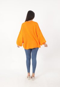 Übergroßer orangefarbener Strickpullover mit breiten, ballonartigen Ärmeln und geripptem Ausschnitt. Kombiniert mit schmal geschnittenen blauen Jeans und durchsichtigen Sandalen mit Absatz.
