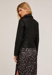 Giacca di pelle nera con un design aderente, collo abbottonato e finitura testurizzata, abbinata a una gonna in pizzo trasparente con motivi intricati.