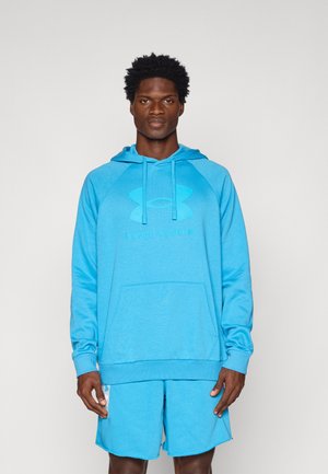 Homme portant un sweat à capuche Under Armour bleu vif avec une poche kangourou et un short assorti, debout devant un fond clair uni.