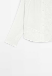 Camicia bianca a maniche lunghe con bottoni sul davanti, caratterizzata da dettagli in pizzo floreale sulla parte inferiore anteriore e una tasca sul petto.