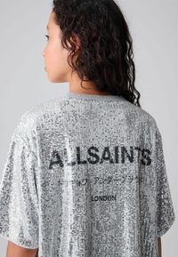 Srebrny t-shirt oversize z cekinami, z żeberkowym dekoltem i czarnym napisem "ALLSAINTS" na plecach, charakteryzujący się teksturowaną, odbijającą powierzchnią.
