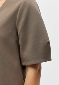 Vêtement marron à manches courtes avec un décolleté en V, fabriqué dans un tissu lisse et texturé. Détail de la couture d'épaule et du bras visible.