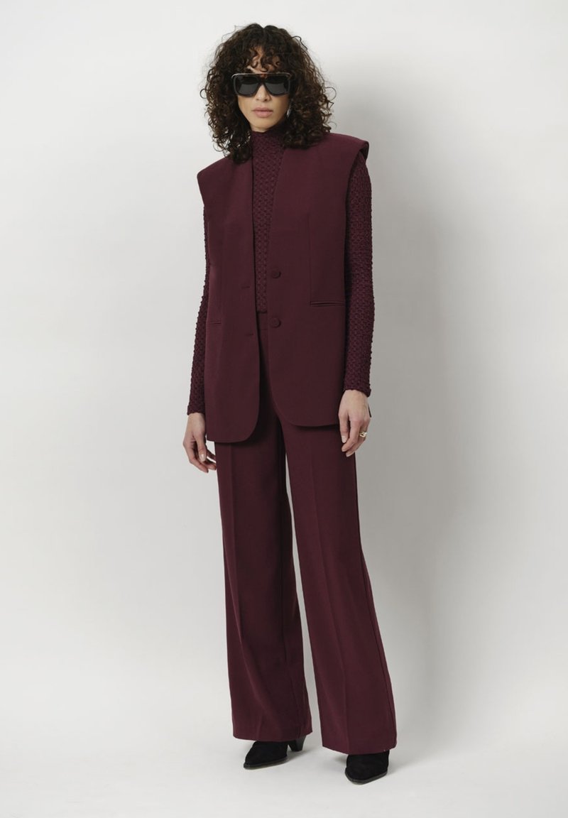 Bordeaux gekleed vest met schoudervullingen en bijpassende brede broek, textuur langwerpig shirt, zwarte vierkante zonnebril en enkellaarsjes.