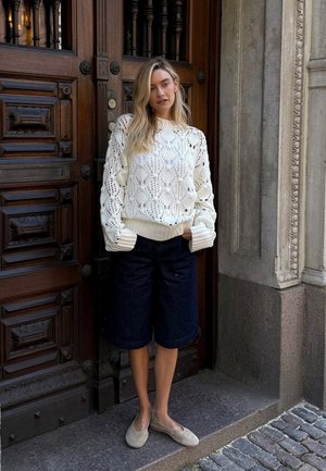 Witte gebreide trui met open patroondessins, crèmekleurige manchetten, gecombineerd met donkere denimshorts en beige instappers, tegen een houten deur.